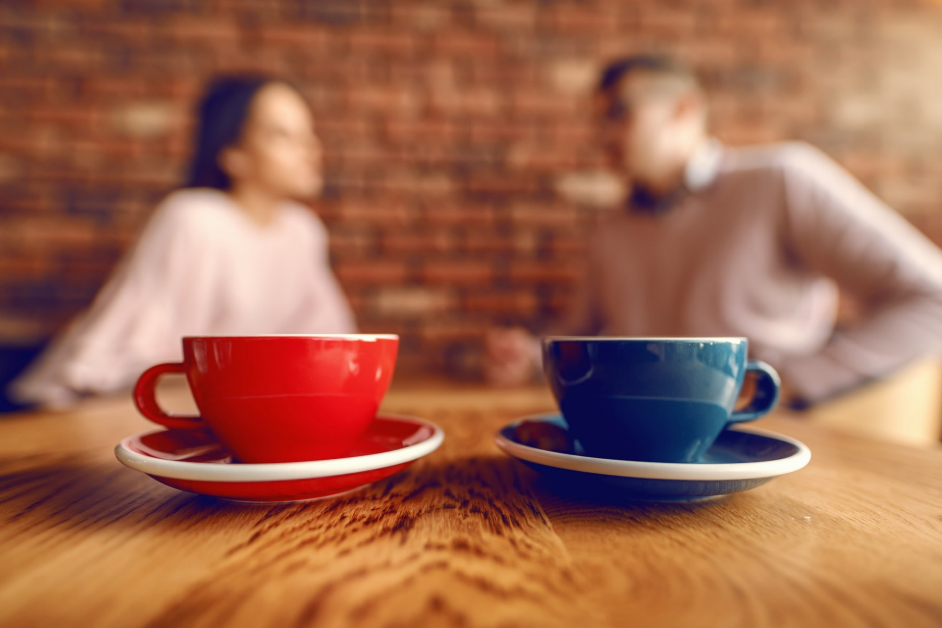 Two cups of coffee in foreground, two people having a conversation in the background.